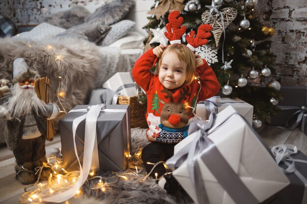 Uma menina com um suéter vermelho quente senta-se sob uma árvore de natal com brinquedos e presentes com chifres na cabeça. infância feliz. atmosfera de feriado de ano novo
