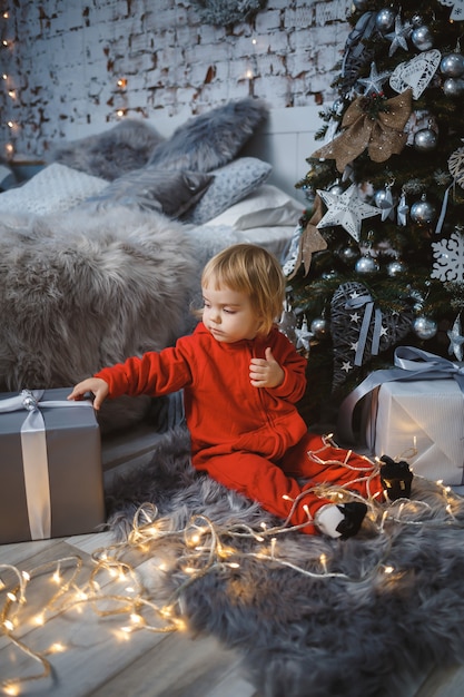 Uma menina com um suéter vermelho quente senta-se sob uma árvore de Natal com brinquedos e presentes. Infância feliz. Atmosfera de feriado de ano novo