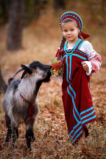 Uma menina com um vestido de verão vermelho ao lado de uma cabra preta na floresta de outono