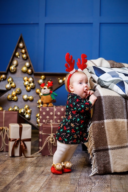 Uma menina com uma camisola quente se senta sob uma árvore de natal com brinquedos e presentes com chifres na cabeça. infância feliz. atmosfera de feriado de ano novo