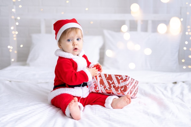 Uma menina com uma fantasia de Papai Noel vermelha está sentada em uma cama com os presentes de Natal em caixas
