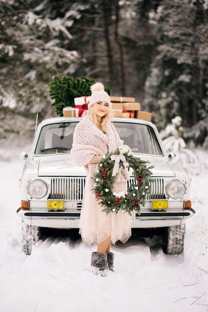 Uma menina com uma guirlanda de Natal nas mãos coberta com um carro retrô em uma floresta de neve