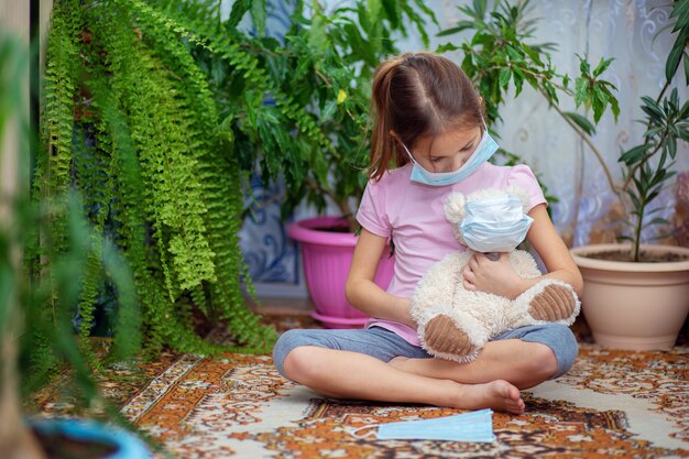 Uma menina com uma máscara médica fica em casa durante uma epidemia de Vieste com seu ursinho de pelúcia também usando uma máscara