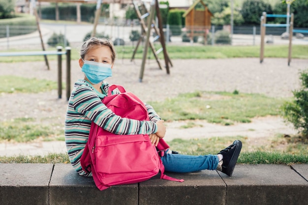 Foto uma menina com uma máscara protetora senta-se no pátio da escola com sua mochila rosa descansando do estudo