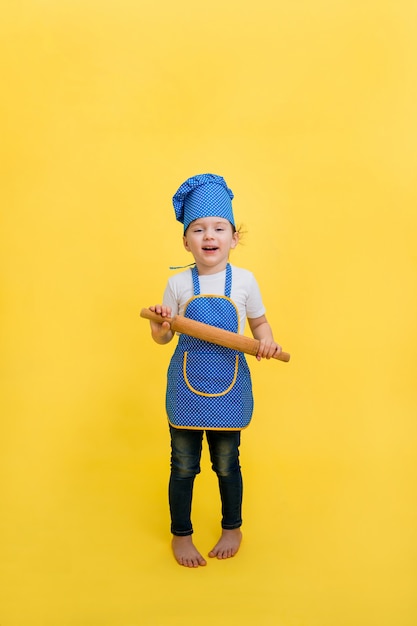 Uma menina cozinha com um rolo em um espaço amarelo com um assento vazio. Uma linda garota de avental e chapéu.