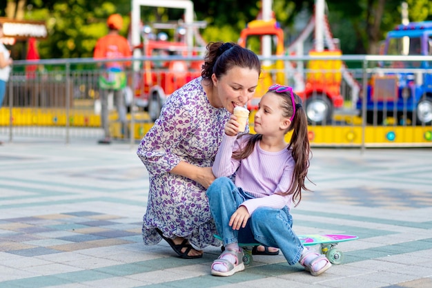 Uma menina criança com sua mãe em um parque de diversões no verão comendo sorvete perto dos carrosséis brincando e rindo do conceito de fins de semana em família e férias escolares