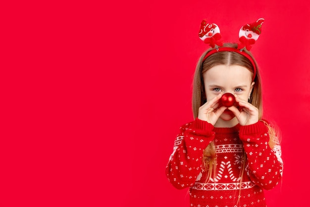 Uma menina criança de suéter com uma bola no nariz em um fundo vermelho monocromático isolado se alegra e sorri o conceito de ano novo e espaço de Natal para texto