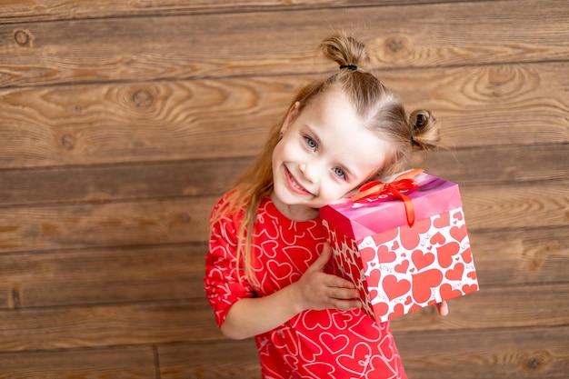 Foto uma menina criança feliz em pijama vermelho com um grande presente nas mãos em um fundo de madeira marrom escuro ri e sorri o conceito de dia dos namorados um lugar para texto