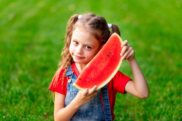 Uma menina criança no verão no gramado com um pedaço de melancia na grama verde se diverte e se alegra mordendo espaço para texto