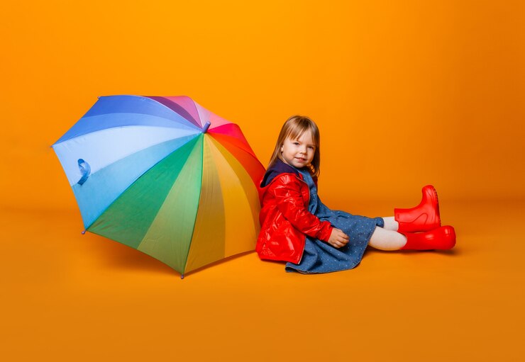 Foto uma menina criança sorridente com uma jaqueta vermelha e botas, segurando um guarda-chuva, fica sobre um fundo amarelo. menina com roupas brilhantes segura um guarda-chuva de arco-íris.