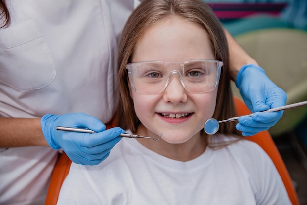 Uma menina de 10 anos com óculos de proteção senta-se sorrindo em uma cadeira odontológica antes de iniciar o tratamento em uma clínica odontológica No quadro as mãos do dentista em luvas azuis com ferramentas