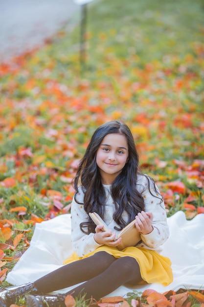 Uma menina de 10 anos de cabelos escuros em um suéter de malha lê um livro sentado em uma clareira em um parque de outono Piquenique na natureza Criança feliz está aprendendo