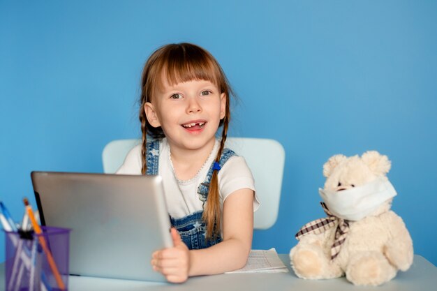 Uma menina de 5 a 6 anos sentada à mesa executa tarefas remotas de lição de casa sobre o tablet. Isolar em uma parede azul.