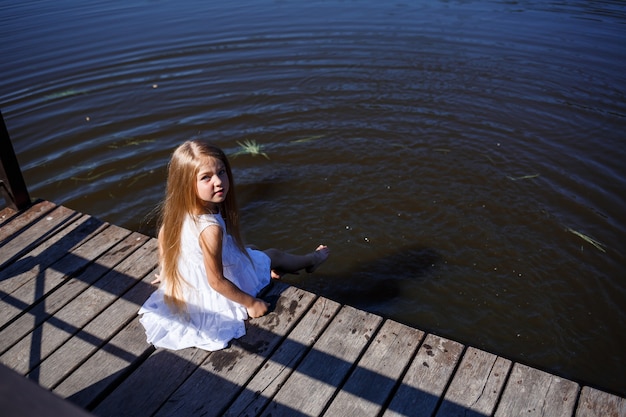 Uma menina de 7 anos com longos cabelos loiros à beira do lago está sentada em uma bolsa com as pernas na água. Ela espirra os pés no lago. Menina descalça em um vestido branco com cabelo comprido.