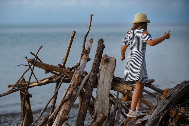 uma menina de 8 anos em um vestido listrado e um chapéu de palha fica em troncos à beira-mar