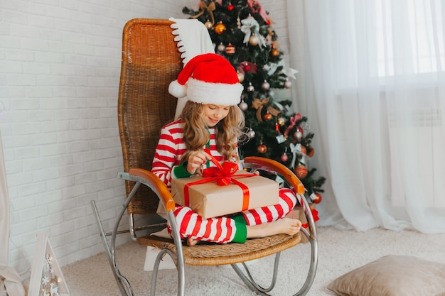 Uma menina de 9 anos segurando um presente do Papai Noel e sentada em uma cadeira de balanço com guirlandas ao fundo. Época de Natal. Feliz Ano Novo!
