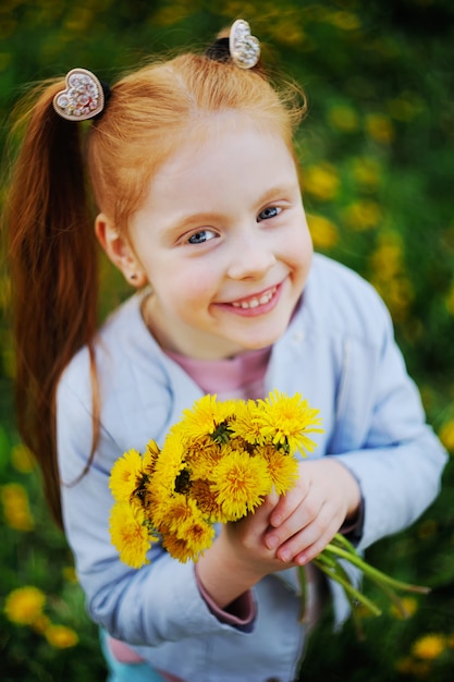 Uma menina de cabelo vermelho sorri contra um campo de dentes de leão e grama verde. Verão, infância, férias