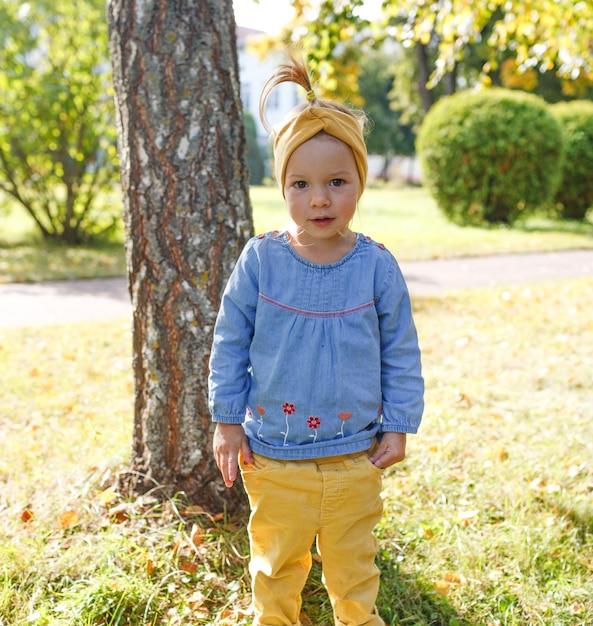 Uma menina de camisa azul e calça amarela está parada no gramado do parque e olha para a câmera
