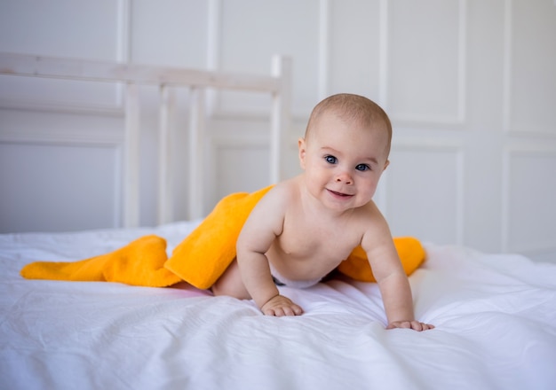Uma menina de fralda e com uma toalha laranja engatinha em um cobertor de algodão branco na cama do quarto
