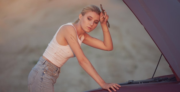 Uma menina de jeans e uma camiseta branca em uma viagem de estrada está reparando um carro em uma estrada de areia