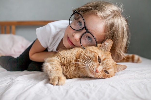 Uma menina de óculos deita na cama e abraça um gato ruivo gordo