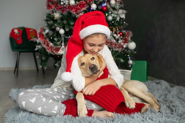 Uma menina de pijama e chapéu de Papai Noel abraça um cachorro labrador perto de uma árvore de Natal no ano novo