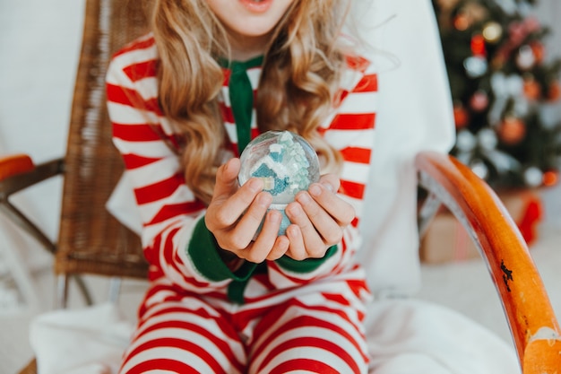 Uma menina de pijama e um chapéu de Natal segura uma bola de Natal - um globo de neve. Natal