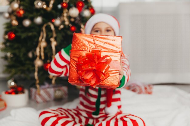 Uma menina de pijama listrado vermelho perto de uma árvore de Natal decorada está segurando um presente de Natal. Conceito de férias de Natal e ano novo.