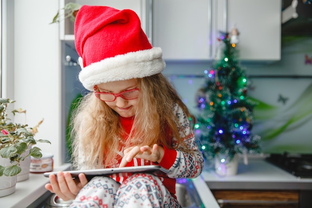 Uma menina de quatro anos com um chapéu de papai noel e óculos escreve uma carta no bloco para o papai noel enquanto está sentada na mesa da cozinha