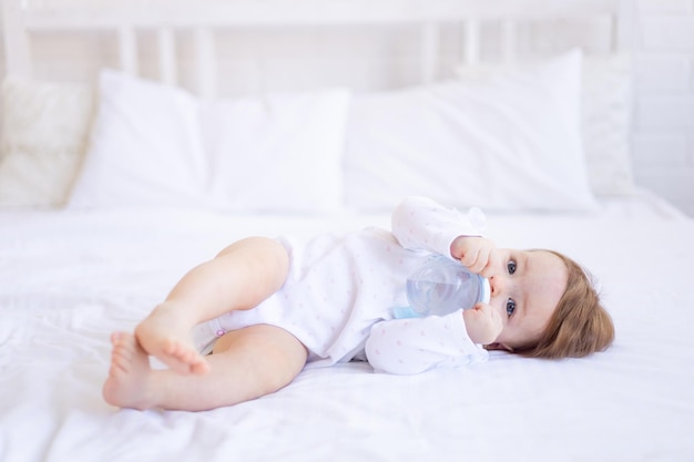 Uma menina de seis meses em uma cama com roupas brancas suga uma garrafa de água ou leite uma criança pequena em uma cama de algodão em casa acordou de manhã comendo o conceito de comida para bebê