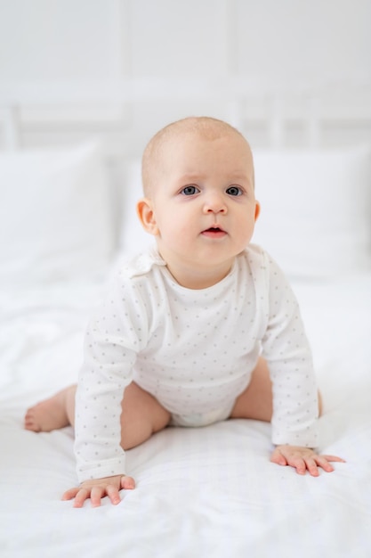 Uma menina de seis meses está sentada em uma cama de algodão branco em um macacão na cama em casa e sorrindo