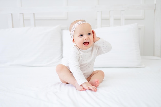 Uma menina de seis meses segura a mão no ouvido em uma cama de algodão branco em casa e sorri