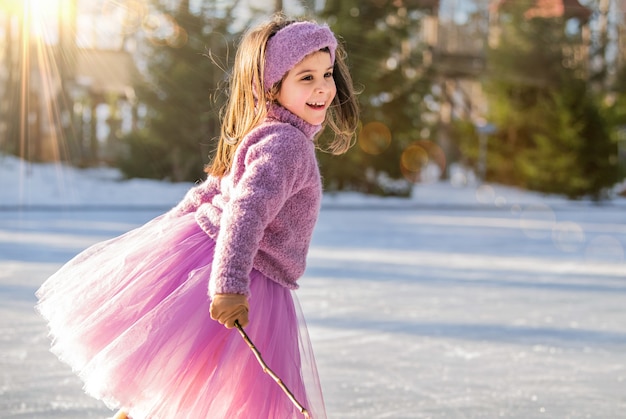 Uma menina de suéter rosa e saia cheia cavalga em um dia ensolarado de inverno em uma pista de gelo ao ar livre no parque