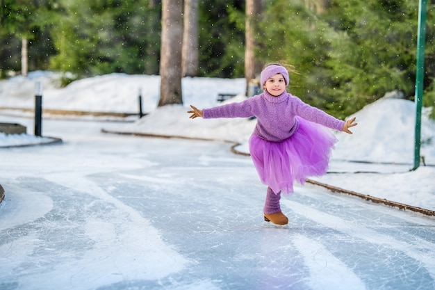 Uma menina de suéter rosa e saia cheia cavalga em um dia ensolarado de inverno em uma pista de gelo ao ar livre no parque