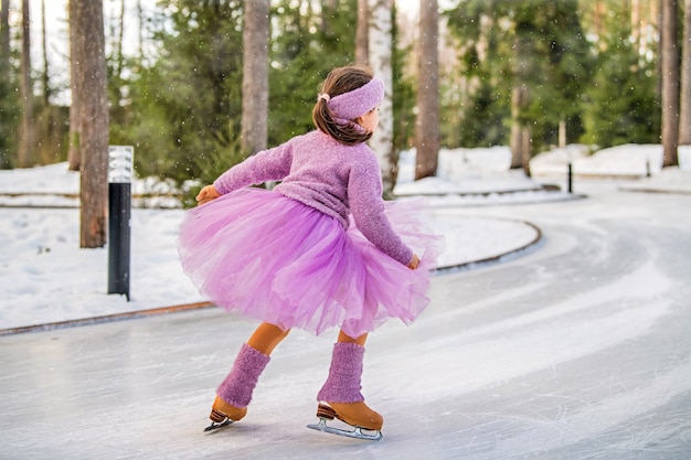 Uma menina de suéter rosa e saia cheia cavalga em um dia ensolarado de inverno em uma pista de gelo ao ar livre no parque