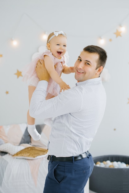 Uma menina de um ano em um vestido rosa está feliz e sorrindo nos braços do pai