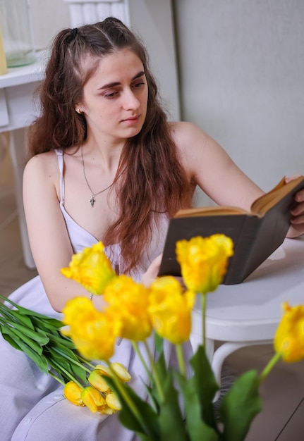 Uma menina de vestido branco e com cabelos longos lê um livro contra um fundo de flores amarelas