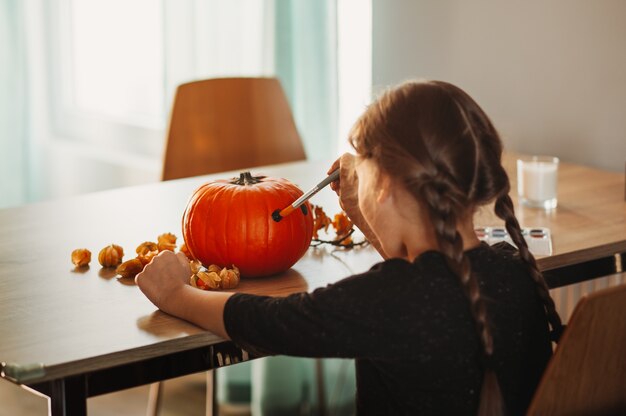 Uma menina decora uma abóbora em uma sala para o Halloween. Crianças decoram a casa. Crianças esculpem abóbora. Divertimento assustador de Halloween