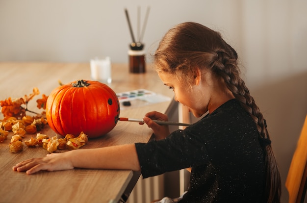 Uma menina decora uma abóbora em uma sala para o Halloween. Crianças decoram a casa. Crianças esculpem abóbora. Divertimento assustador de Halloween