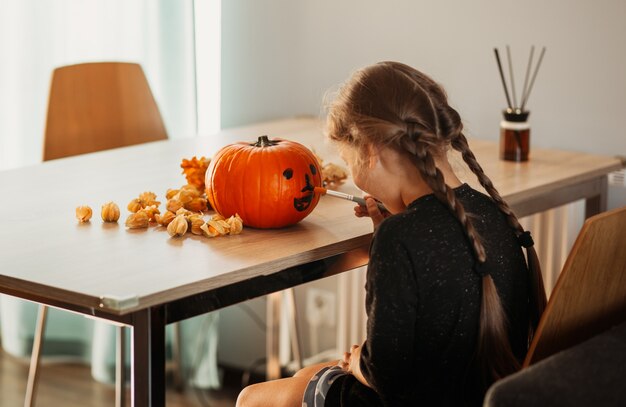 Uma menina decora uma abóbora em uma sala para o Halloween. Crianças decoram a casa. Crianças esculpem abóbora. Divertimento assustador de Halloween