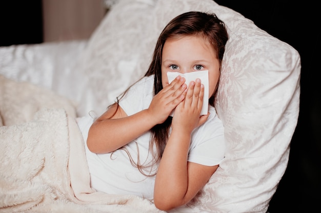 Uma menina deita na cama em um lindo quarto, espirra e cobre o nariz com um lenço, o frio do outono, a segunda onda do vírus