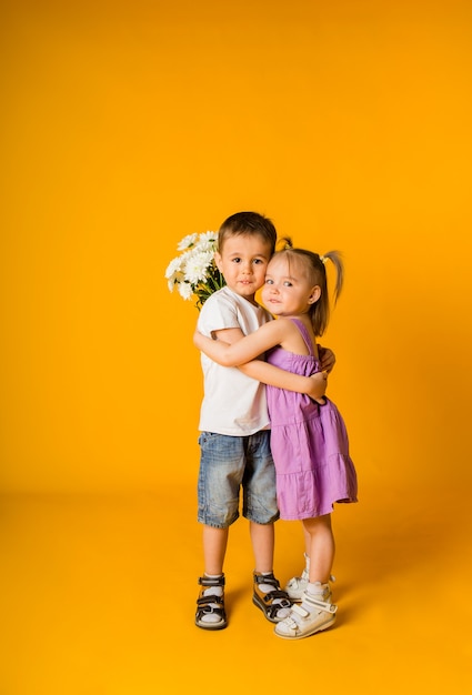 Uma menina e um menino se abraçam e seguram um buquê de flores em uma superfície amarela com espaço para texto