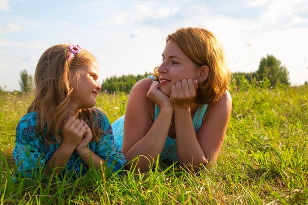 Uma menina e uma mulher-mãe deitam-se na grama verde em um campo, o tempo ensolarado de verão, o sorriso e a alegria de uma criança, uma filha conta os segredos de seus filhos