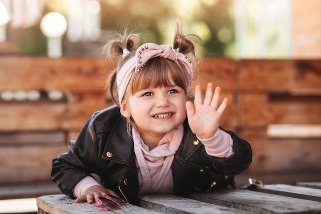 Uma menina elegante com uma jaqueta de couro preta senta e ri no terraço de um café de verão