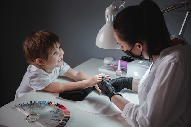 Uma menina em um salão de beleza. Olha a manicure em uma máscara preta e luvas pretas. Cuidar das unhas da criança.