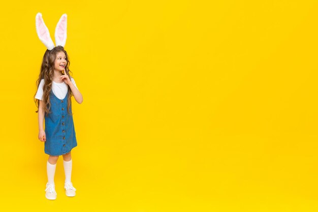Uma menina em um vestido azul com orelhas de coelho fica em um fundo amarelo.