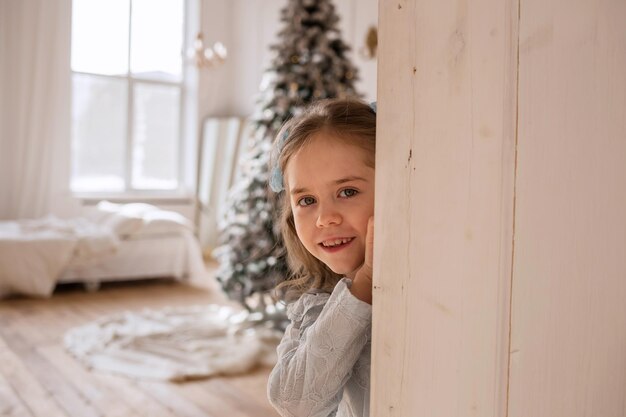 Uma menina em um vestido de renda azul está brincando de esconde-esconde atrás de uma porta branca em um quarto