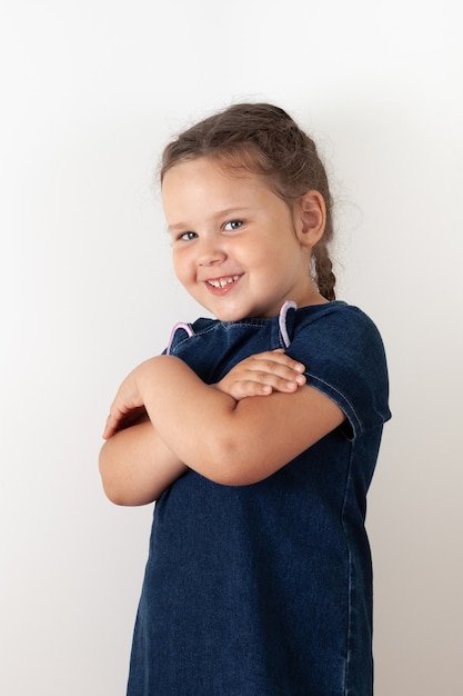 Uma menina em um vestido jeans azul tem os braços cruzados sobre o peito e está de lado. Criança alegre, isolada.