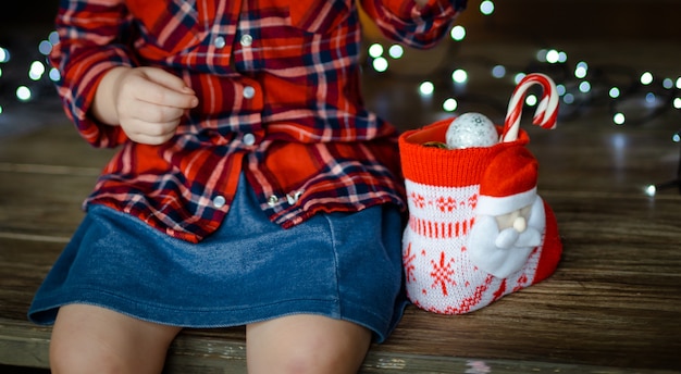 Uma menina em uma camisa xadrez vermelha e uma saia jeans azul, abre os doces do presente de Natal, sentado em uma mesa de madeira. Conceito de manhã de Natal. Fechar-se.