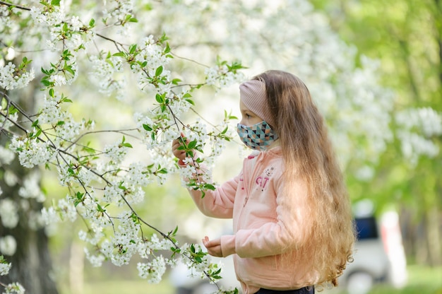 Uma menina em uma máscara médica olha para uma flor de árvore.